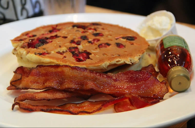 Stack of fresh cranberry pancakes with whipped butter, bottle of pure maple syrup and a side of bacon